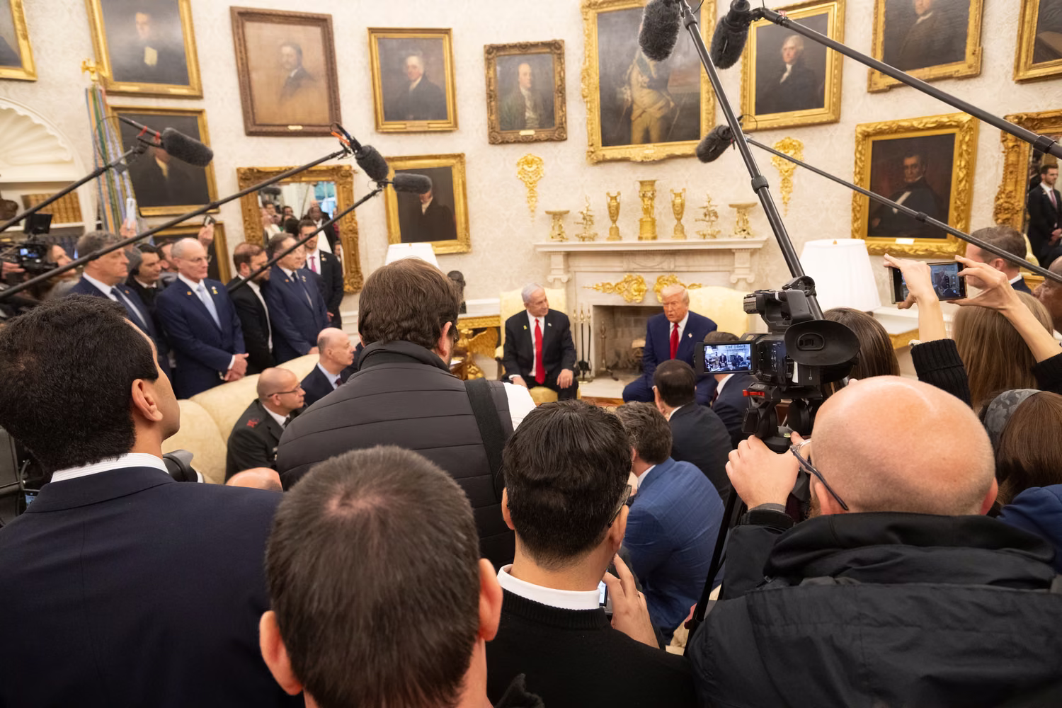 Two men sit at table in front of scrum of cameramen and reporters seen from behind.