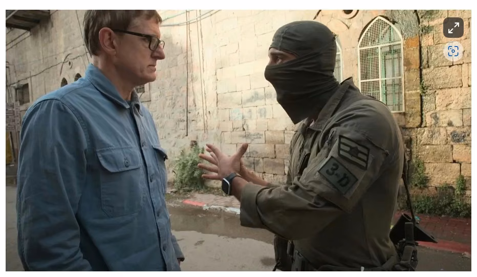 Bespectacled white male in blue shirt on left faces a masked soldier