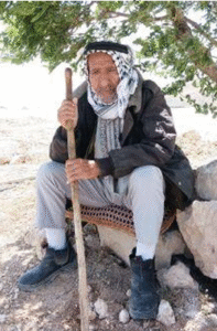 Old Palestinian man seated outside with a walking stick in his hands, a keffiyeh on his head