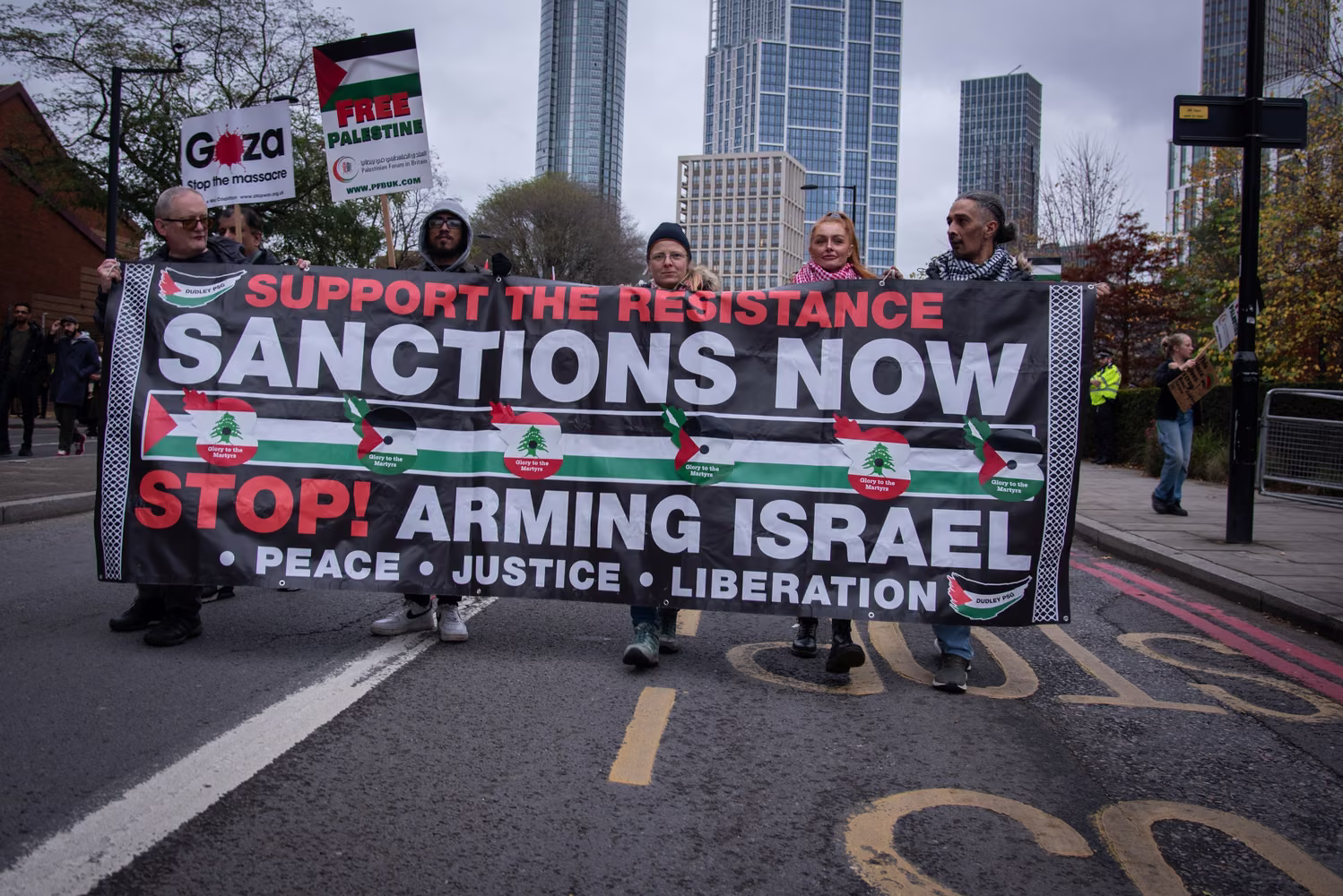 Lrge banner statiung "Sanctions Now. Stop Arming Israel" on march for Palestine. Demonstrations can be seen behind the large banner
