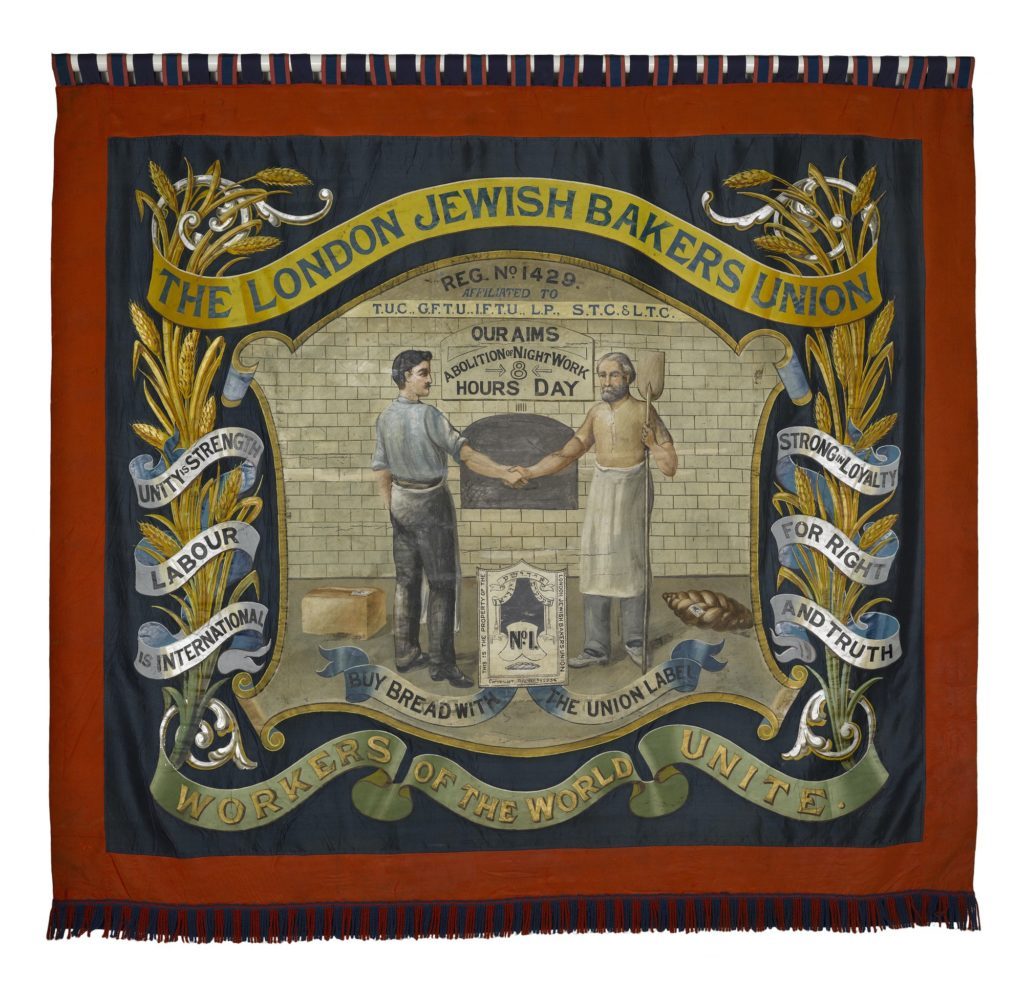 ornate embroidered banner, two men shaking hands in centre, sheaves of wheat entwined with slogans right and left